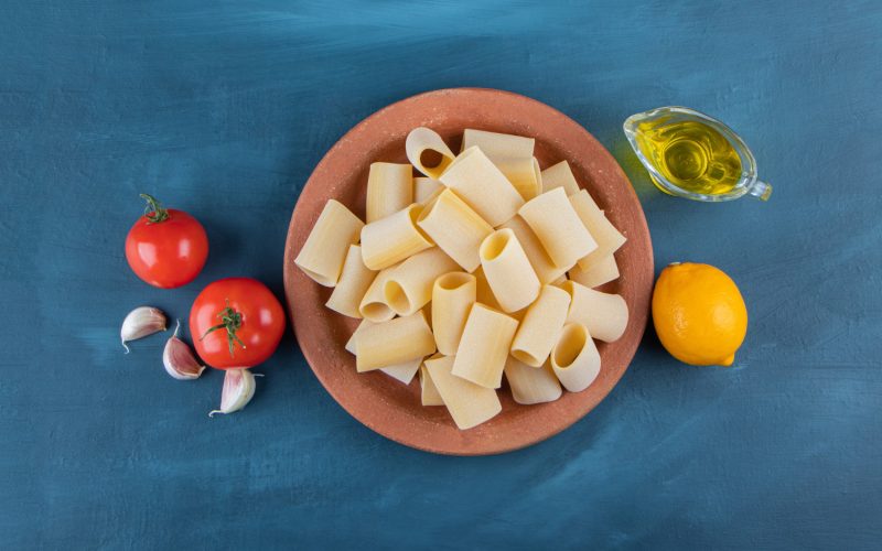 A brown plate of raw cannelloni pasta with fresh red tomatoes and lemon on a dark-blue background